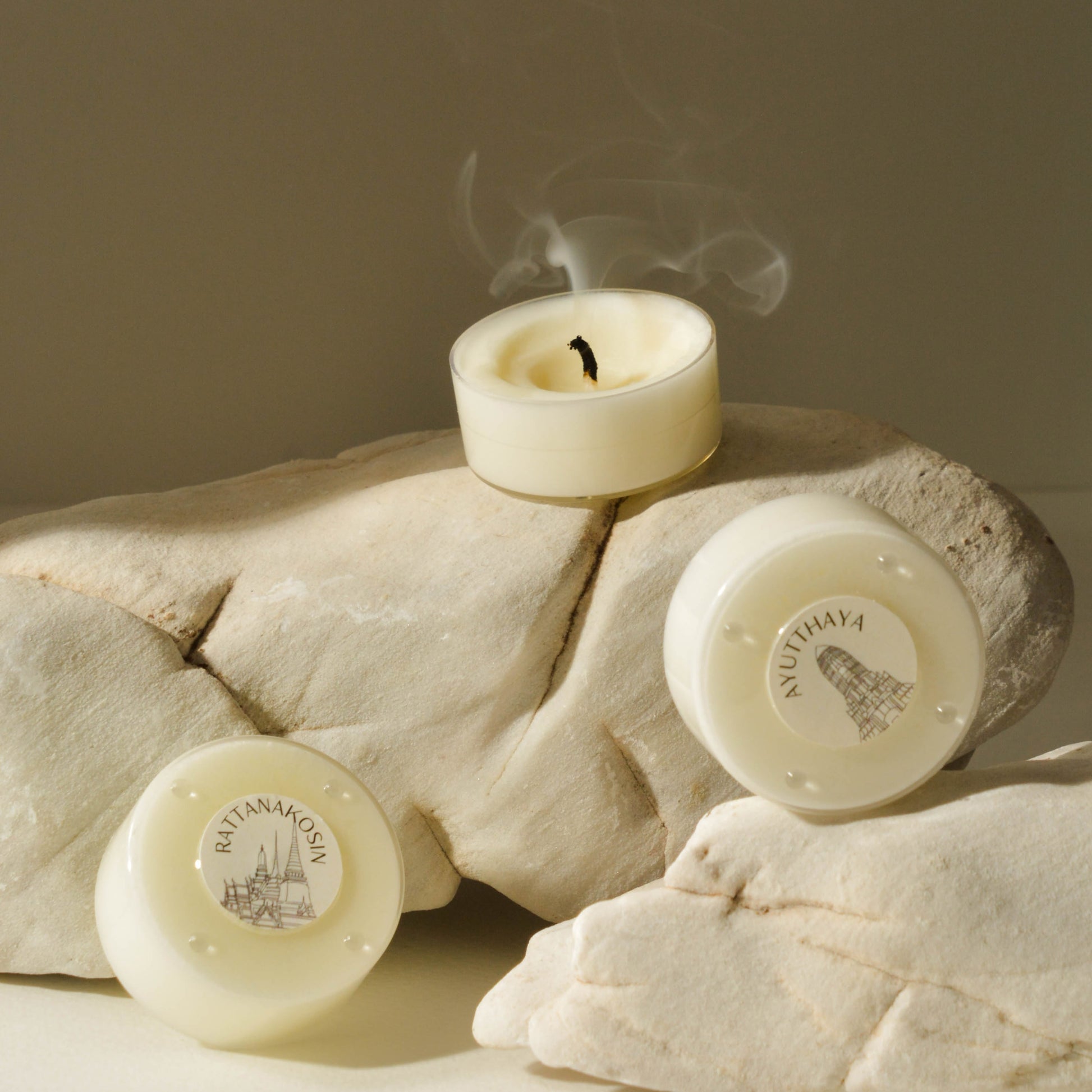 A beige candle with a white label placed on a rock, with a visible wick and smoke rising from the top.