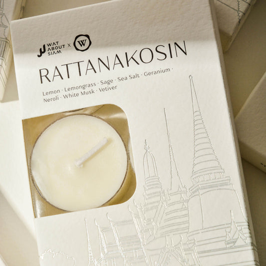 A hand-poured tealight candle is displayed in an open box with 'RATTANAKOSIN' printed on it. The candle is white and appears to be made of a natural wax, and the box has an illustration of a Thai temple.