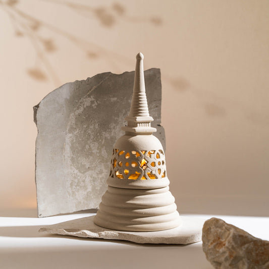 A matte white ceramic candle holder designed in the shape of a Sukhothai-style Stupa with a stencil pattern, displayed against a neutral background with a rock and a shadow cast upon it.
